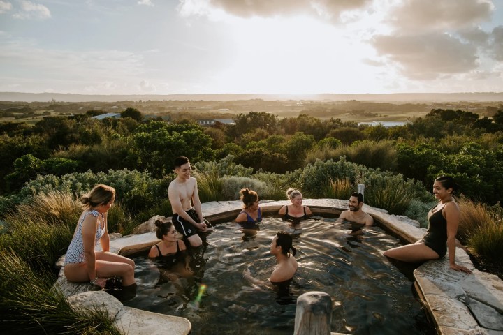 Group of people enjoying a hot spring with a scenic landscape view at sunset.